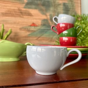 May include: A light gray teacup with a curved handle sits on a wooden surface. Behind it, a stack of colorful teacups in red, green, and gray are visible. A green serving bowl and plates are also in the image, suggesting a tea set.