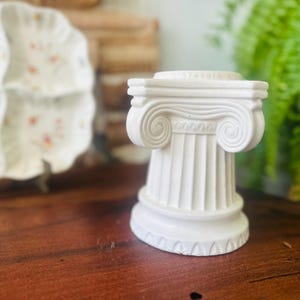 May include: A small, white, decorative pillar in the classical Ionic style. The pillar has a fluted column and a detailed capital with scroll-like volutes. The object is made of a solid material and sits on a dark wooden surface.