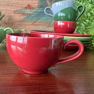 May include: A red ceramic teacup with a curved handle sits on a wooden surface. Behind it, a stack of teacups in red, green, and gray are visible, along with matching saucers. A green plant and a decorative backdrop complete the scene.