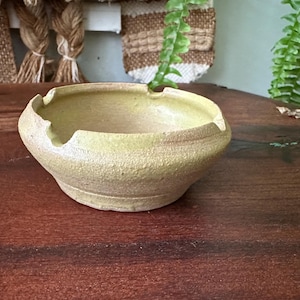 May include: A small, round, handmade ceramic ashtray with a textured surface and a light yellow-green color. The ashtray has a slightly irregular rim with notches. It sits on a dark brown wooden surface.