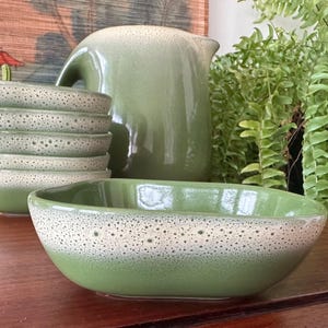 May include: A green ceramic serving bowl with a speckled white rim. A stack of matching bowls, a pitcher, and a fern are in the background. The ceramic pieces have a two-tone glaze.