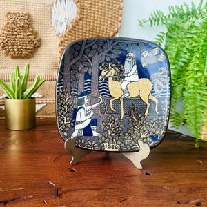May include: Square decorative plate with a dark blue background and a gold and white illustration of a person riding a horse and another person with a bow and arrow. The plate is on a gold stand.