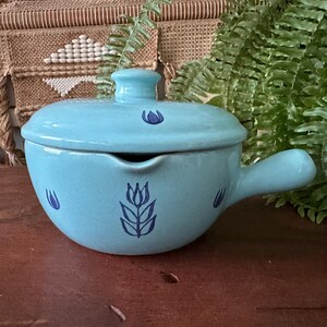 May include: A light blue ceramic soup bowl with a lid and handle. The bowl is decorated with dark blue floral designs. The bowl is on a wooden surface.