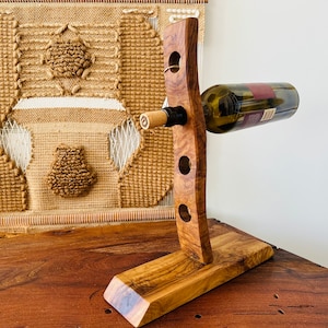May include: A wooden wine bottle holder with a curved design and a base. The holder has four holes to hold bottles and is made of light brown wood.