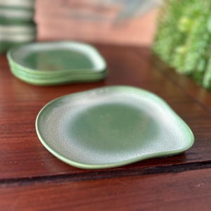 May include: A set of green ceramic plates with a unique, organic shape. The plates have a speckled, textured surface and a slightly raised edge. The plates are stacked on a dark wooden surface, with a blurred background.