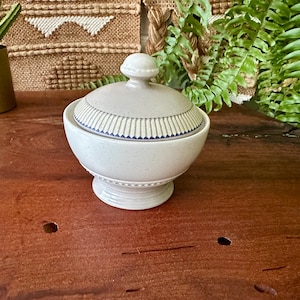 May include: A white ceramic sugar bowl with a lid, featuring a pedestal base and a decorative blue and white striped pattern around the rim. The bowl is on a wooden surface.