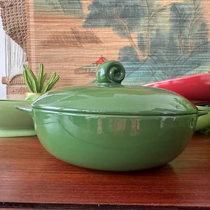May include: A green ceramic serving bowl with a lid, featuring a spiral handle. A matching green soup bowl with a handle is visible to the left. A red serving dish is in the background. The items are on a wooden surface.