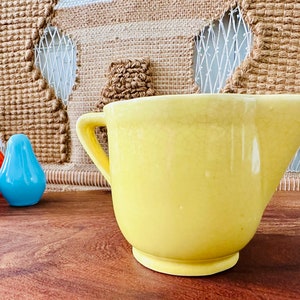 May include: A yellow ceramic pitcher with a handle. The pitcher has a textured surface and is sitting on a wooden table.