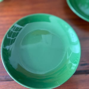 May include: A close-up shot of a bright green ceramic bowl. The bowl has a glossy finish and a rounded shape. The bowl is sitting on a wooden surface. Another bowl is partially visible in the background.