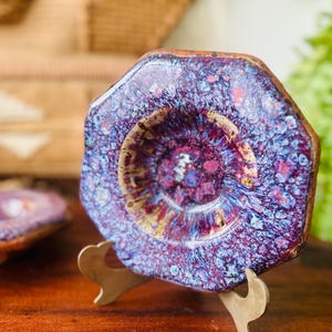 May include: A ceramic plate with a unique octagonal shape and a vibrant, speckled glaze in shades of purple, blue, and red. The plate is displayed on a gold-colored stand, showcasing its artistic design. The plate is a decorative item.