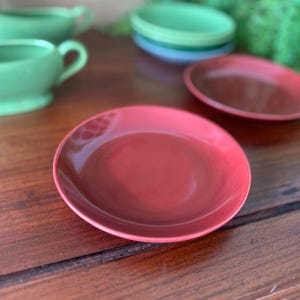 May include: A collection of ceramic dishware in shades of red and green. The set includes a red plate, a green soup bowl with a handle, and other bowls. The items are arranged on a wooden surface.
