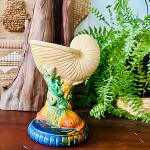 May include: A decorative ceramic nautilus shell-shaped vase. The vase is beige with a spiral design, supported by a colorful base featuring fish and aquatic plants. The base is blue, green, orange, and yellow. The vase is a unique home decor item.