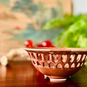May include: A brown ceramic bowl featuring a decorative lattice cutout pattern around the upper rim. The bowl has a smooth, glazed finish and a rounded base, showcasing a consistent reddish-brown tone throughout the exterior surface.