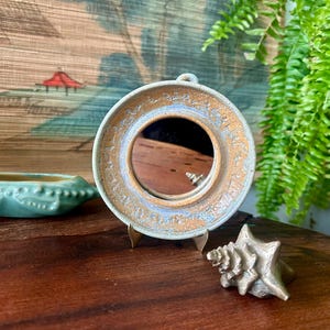 May include: A small, round, ceramic mirror with a light brown and blue rim, displayed on a wooden stand. A small, turquoise ceramic dish and a silver seashell are also on the wooden surface.