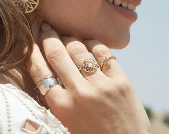 May include: A close-up of a hand wearing three gold rings. The ring on the index finger features a delicate, circular design with a small, clear stone in the centre. The ring on the middle finger is a simple, thin band. The ring on the ring finger is a wider band with a hammered texture.
