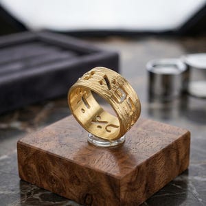 May include: A wide gold band ring featuring a textured, brushed finish on the exterior surface. The ring is designed with cut-out Hebrew characters that wrap around the entire circumference of the band, creating an openwork pattern throughout.