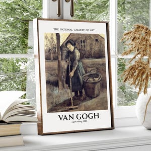 Op de afbeelding: Een ingelijste prent van Van Goghs "A Girl Raking" van The National Gallery of Art. Het kunstwerk toont een vrouw die aan het harken is, met een emmer in de buurt. De prent wordt tentoongesteld op een witte vensterbank met een vaas en een open boek.