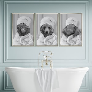 May include: Three black and white photographs of animals wearing white bathrobes. The animals are a cow, a bear, and an elephant. The photographs are framed and hanging on a pale blue wall above a white bathtub.