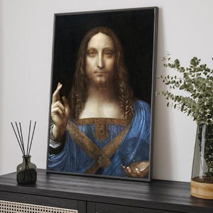 May include: Framed print of a Renaissance portrait. The subject, with long brown hair and a beard, wears a blue robe with gold detailing. The artwork is displayed on a dark surface, with a vase of greenery and a diffuser nearby.