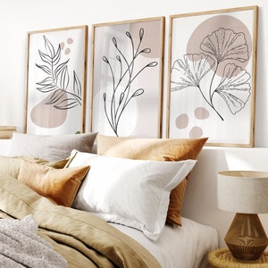 Könnte beinhalten: Drei gerahmte Drucke mit minimalistischen Linienzeichnungen von Blättern und Zweigen auf hellem Hintergrund mit dezenten rosafarbenen Akzenten. Die Drucke hängen an einer weißen Wand über einem Bett mit brauner Bettwäsche und Kissen.