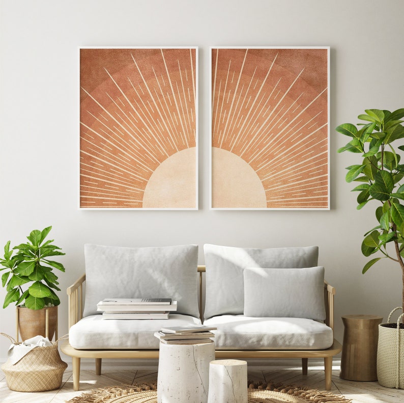 Op de afbeelding: Twee ingelijste prints van een gestileerde zon met oranje en witte stralen op een bruine achtergrond. De prints hangen aan een witte muur boven een lichtgrijze bank met twee kussens.