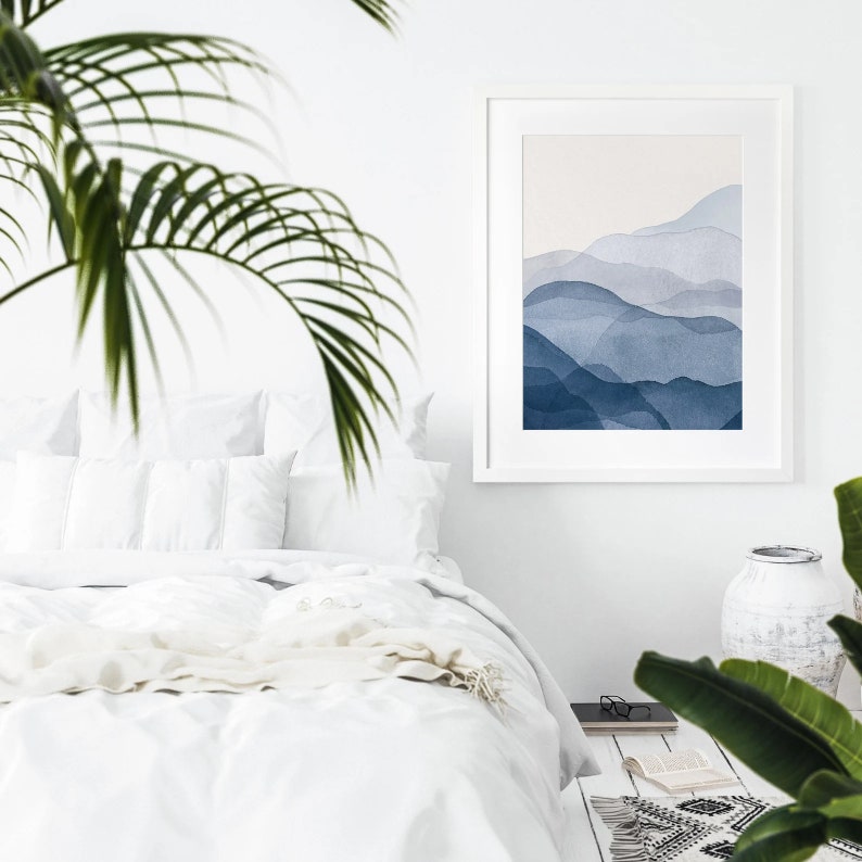May include: A white framed print of a watercolor painting of blue mountains. The print is hanging on a white wall above a white bed with white bedding and a white throw blanket.