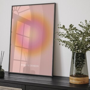 May include: A framed print with a pink and orange gradient background and the text "POSITIV DENKEN" in black. The print is hanging on a white wall above a black table with a vase of green eucalyptus.
