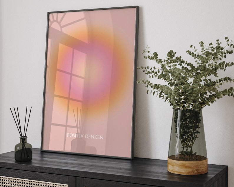 May include: A framed print with a pink and orange gradient background and the text "POSITIV DENKEN" in black. The print is hanging on a white wall above a black table with a vase of green eucalyptus.