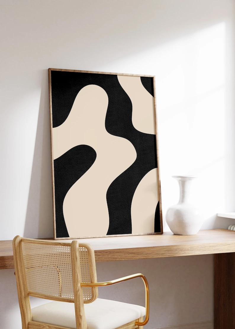 May include: A framed abstract print with a black and beige color scheme. The print features a series of organic shapes that resemble waves or clouds. The print is hanging on a white wall above a wooden desk with a chair.