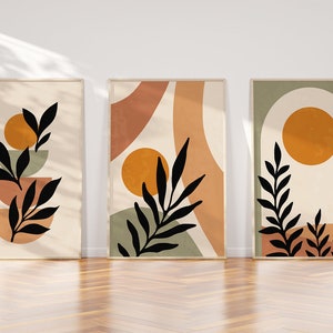 Op de afbeelding: Drie abstracte kunstprints met een minimalistisch ontwerp met zwarte silhouetten van bladeren en takken tegen een achtergrond van beige, oranje, groene en bruine geometrische vormen.