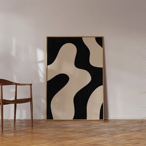 May include: A black and white abstract art print with organic shapes in a wooden frame. The print is hanging on a white wall in a room with a wooden floor and a chair.