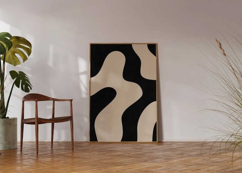 May include: A black and white abstract art print with organic shapes in a wooden frame. The print is hanging on a white wall in a room with a wooden floor and a chair.