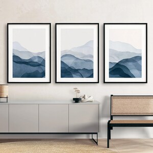 May include: Three framed prints of abstract watercolor paintings depicting blue and white mountains. The prints are hung on a white wall above a gray cabinet.
