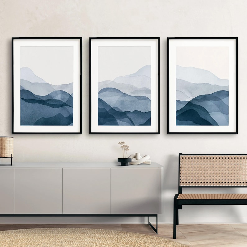 May include: Three framed prints of abstract watercolor paintings depicting blue and white mountains. The prints are hung on a white wall above a gray cabinet.