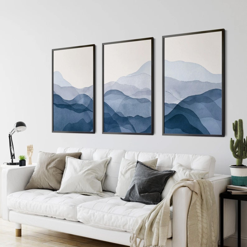 May include: Three framed abstract watercolor paintings with a blue and white color scheme depicting a mountain range. The paintings are hung on a white wall above a white couch with pillows.