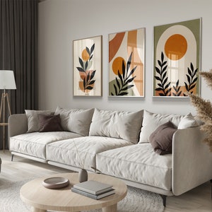 Op de afbeelding: Drie abstracte kunstprints in een moderne minimalistische stijl. De prints bevatten geometrische vormen, bladeren en cirkels in tinten oranje, bruin en groen. De prints zijn ingelijst in een licht houten lijst en hangen aan een witte muur.