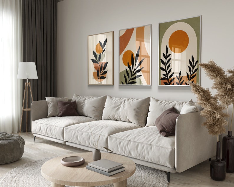Op de afbeelding: Drie abstracte kunstprints in een moderne minimalistische stijl. De prints bevatten geometrische vormen, bladeren en cirkels in tinten oranje, bruin en groen. De prints zijn ingelijst in een licht houten lijst en hangen aan een witte muur.