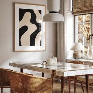 May include: A dining room table with a white marble top and gold accents. The table is surrounded by four chairs with woven cane seats and backs. A white ceramic pendant light hangs above the table. A framed abstract print with black and beige shapes hangs on the wall behind the table.