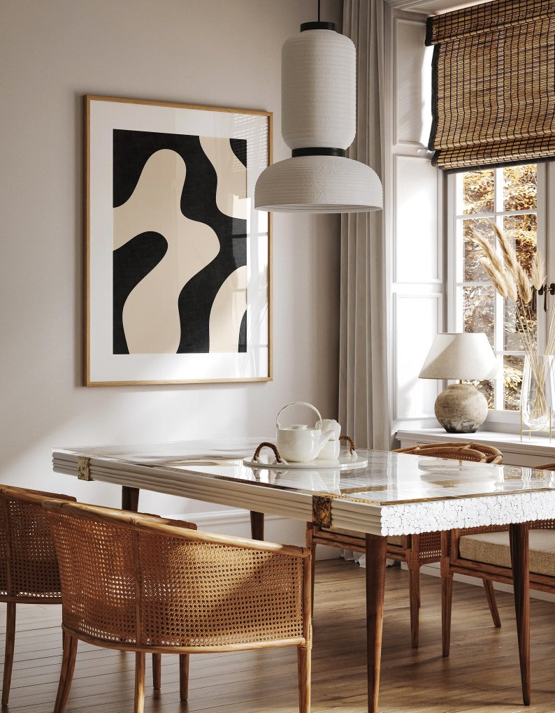 May include: A dining room table with a white marble top and gold accents. The table is surrounded by four chairs with woven cane seats and backs. A white ceramic pendant light hangs above the table. A framed abstract print with black and beige shapes hangs on the wall behind the table.