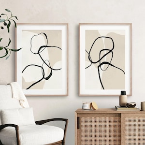May include: Two framed abstract art prints with black line drawings on a beige background. The prints are in simple wooden frames and are hanging on a white wall.
