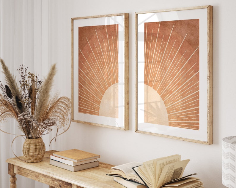 Op de afbeelding: Twee ingelijste prints van een gestileerde zon in oranje en bruine tinten. De zon heeft een wit centrum en stralende lijnen. De prints hangen aan een witte muur boven een houten tafel met boeken.