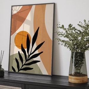 Op de afbeelding: Abstracte kunstprint met een zwart silhouet van een blad, oranje en bruine vormen en een witte achtergrond. De print is ingelijst in een zwart frame en hangt aan een witte muur.