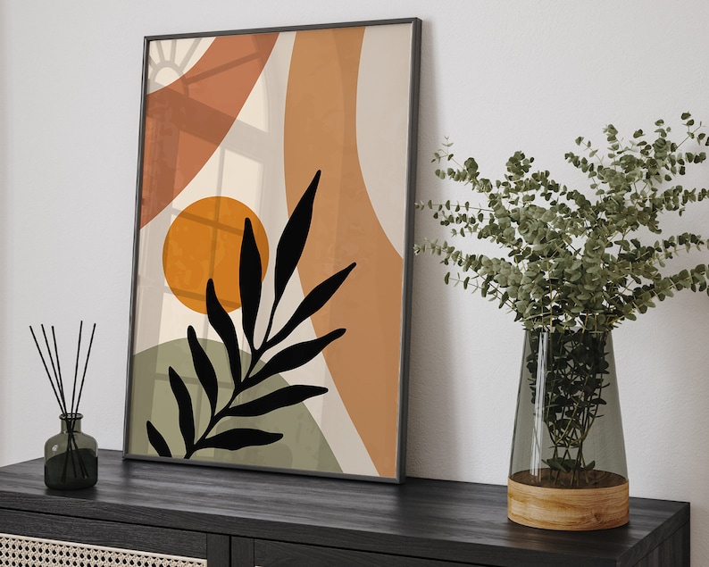 Op de afbeelding: Abstracte kunstprint met een zwart silhouet van een blad, oranje en bruine vormen en een witte achtergrond. De print is ingelijst in een zwart frame en hangt aan een witte muur.