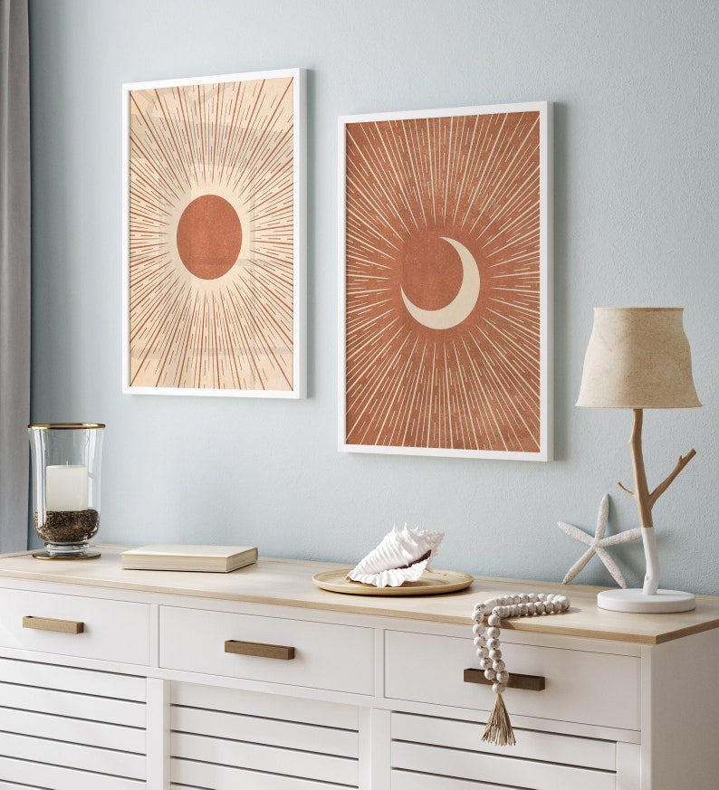 Puede incluir: Dos impresiones enmarcadas con un dise&ntilde;o minimalista que presenta un sol y una luna creciente. Las impresiones tienen un marco blanco y un fondo beige con formas de sol y luna naranja. Las impresiones est&aacute;n colgadas en una pared azul claro sobre una c&oacute;moda blanca.
