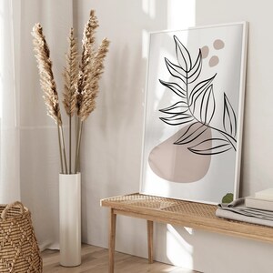 Peut inclure: Une impression encadr&eacute;e blanche avec un dessin au trait noir d'une feuille avec une forme abstraite rose. L'impression est accroch&eacute;e &agrave; un mur blanc au-dessus d'un banc en bois clair avec un si&egrave;ge tiss&eacute;.