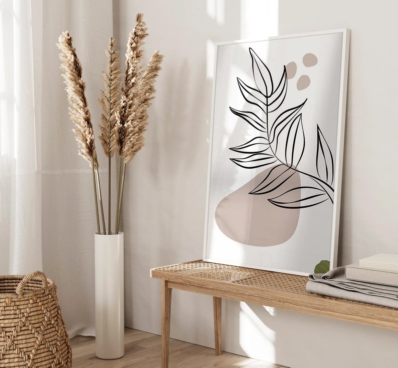 Peut inclure: Une impression encadr&eacute;e blanche avec un dessin au trait noir d'une feuille avec une forme abstraite rose. L'impression est accroch&eacute;e &agrave; un mur blanc au-dessus d'un banc en bois clair avec un si&egrave;ge tiss&eacute;.