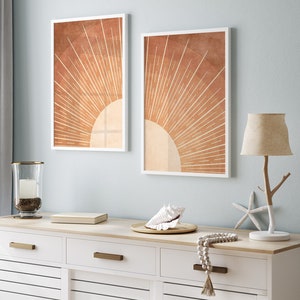 Op de afbeelding: Twee ingelijste prints van een gestileerde zon met oranje en bruine stralen op een lichtblauwe muur boven een witte commode met een schelp, een lamp en een kralenketting.
