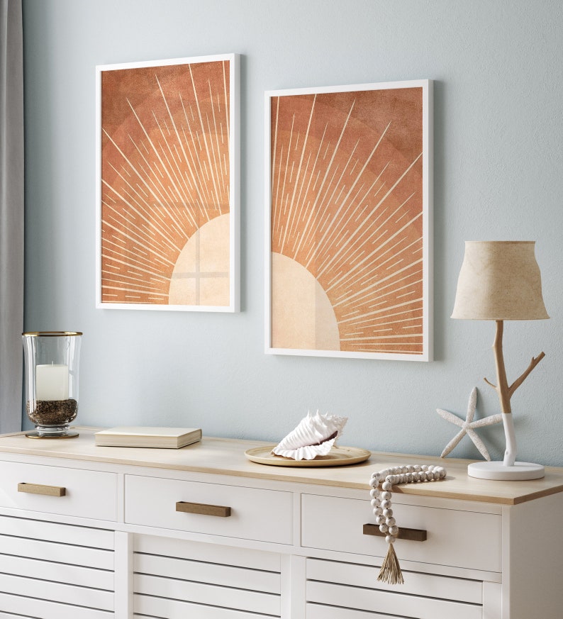 Op de afbeelding: Twee ingelijste prints van een gestileerde zon met oranje en bruine stralen op een lichtblauwe muur boven een witte commode met een schelp, een lamp en een kralenketting.