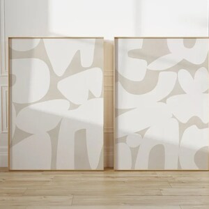 Op de afbeelding: Twee beige abstracte kunstprints met witte organische vormen in een minimalistische stijl. De prints zijn ingelijst in een dun gouden frame en hangen aan een witte muur.