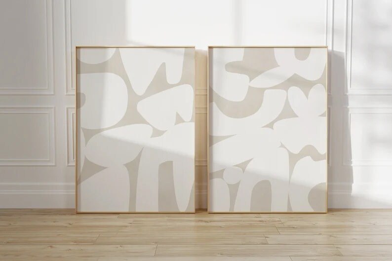 Op de afbeelding: Twee beige abstracte kunstprints met witte organische vormen in een minimalistische stijl. De prints zijn ingelijst in een dun gouden frame en hangen aan een witte muur.